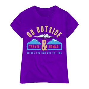 May include: A purple t-shirt with the words "GO OUTSIDE" in gold lettering above a graphic of two blue mountains and the words "TRAVEL & NOMAD" in blue boxes. The text "BEFORE YOU RUN OUT OF TIME" is below the graphic.