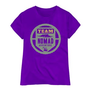 May include: A purple t-shirt with a crew neck and short sleeves. The shirt features a circular graphic with the words "TEAM NOMAD ADVENTURE" in a combination of purple, yellow, and green. The graphic includes cloud and grass illustrations.