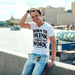 以下が含まれることがあります： 「BORN TO DRINK FORCE TO WORK」の黒い文字がプリントされた白いVネックTシャツを着た人。その人は、水色のジーンズとサングラスをかけています。背景には川、建物、橋があります。