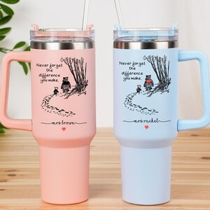 May include: Two insulated tumblers, one pink and one light blue, with handles and straws. Each features a whimsical illustration of a bear and a smaller animal walking through a forest, with the text "Never forget the difference you make."