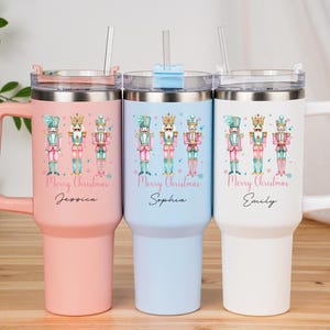 May include: Three insulated tumblers with handles, each featuring a different color: pink, light blue, and white. Each tumbler has a lid, a straw, and a design of three nutcracker figures with the text "Merry Christmas" and a name.