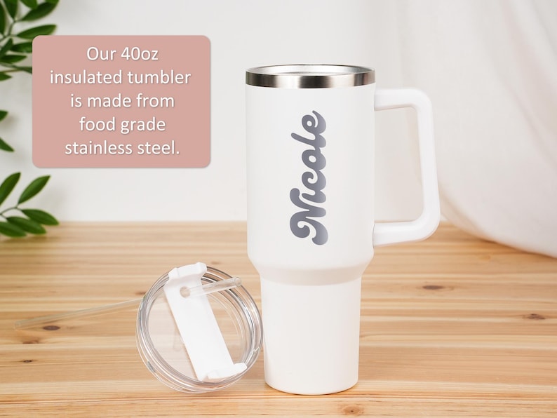 May include: White 40oz insulated tumbler with a handle and a silver rim. The name "Nicole" is printed in gray script on the side. The lid and straw are visible. The text on the image says the tumbler is made from food grade stainless steel.
