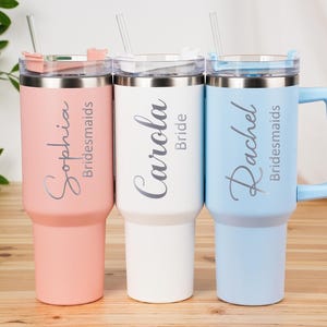 May include: Three insulated tumblers in pastel colors: pink, white, and blue. Each tumbler has a silver lid and straw. The pink tumbler says "Sophia Bridesmaids", the white says "Carola Bride", and the blue says "Rachel Bridesmaids".