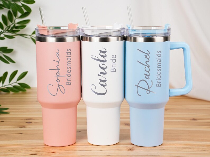 May include: Three insulated tumblers in pastel colors: pink, white, and blue. Each tumbler has a silver lid and straw. The pink tumbler says "Sophia Bridesmaids", the white says "Carola Bride", and the blue says "Rachel Bridesmaids".