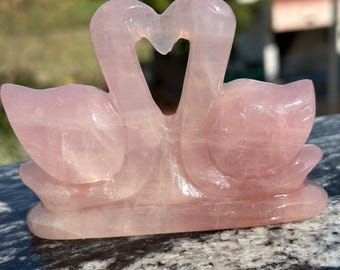 May include: A pink rose quartz sculpture of two swans forming a heart shape. The swans are carved from a translucent, light pink stone and rest on a rounded base. The sculpture is displayed outdoors in natural light.