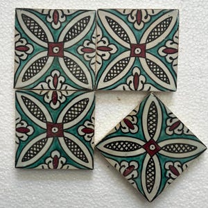 Op de afbeelding: Vier vierkante keramische tegels en één ruitvormige tegel met een herhalend bloemmotief. Het ontwerp heeft turquoise, witte en bordeauxrode kleuren met een geometrisch rooster en bloemelementen. De tegels zijn op een wit oppervlak geplaatst.
