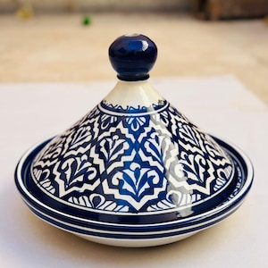 Peut inclure: Un tajine en céramique blanc avec un motif floral bleu. Le couvercle conique est surmonté d'un bouton bleu foncé. Le bord et la base sont bleu foncé. Idéal pour la cuisine marocaine.