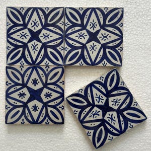 Op de afbeelding: Vier vierkante keramische tegels met een witte achtergrond en een blauw bloemmotief. Het ontwerp heeft een centrale stervorm omgeven door gestileerde bladeren en andere geometrische elementen. Eén tegel is onder een hoek gedraaid.
