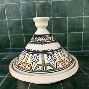 May include: A cream-colored ceramic tagine pot with a conical lid and a round base. The lid is decorated with black, yellow, and green geometric patterns. The tagine is designed for cooking and serving Moroccan dishes.