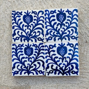 Pode incluir: Quatro azulejos quadrados de cerâmica com fundo branco e design floral azul. Cada azulejo apresenta um motivo central semelhante a uma romã, rodeado por folhas e vinhas estilizadas. Os azulejos estão dispostos em forma quadrada.