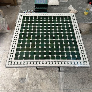 May include: A square table top featuring a mosaic pattern of dark green octagonal tiles separated by small white square tiles. The border consists of a repeating white and black diamond pattern, creating a geometric design on the flat surface.