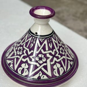 Puede incluir: Una olla de tagine de cerámica morada y blanca con tapa cónica. La tapa y la base están adornadas con un patrón floral geométrico. El borde y la parte superior de la tapa son de color morado liso. Un recipiente de cocina tradicional del norte de África.