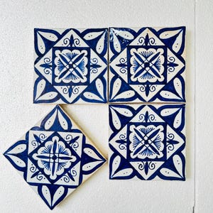 Puede incluir: Cuatro baldosas cerámicas cuadradas y una en forma de diamante con un diseño floral azul y blanco. Las baldosas presentan un motivo floral central rodeado de hojas y espirales estilizadas, creando un patrón decorativo.
