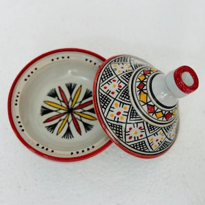 Può includere: Un tagine in ceramica con coperchio, con bordo rosso e intricati motivi geometrici in nero, giallo e rosso. L'interno della pentola presenta un motivo floreale. Il coperchio ha un manico rosso.