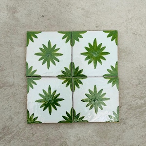 Può includere: Quattro piastrelle quadrate in ceramica bianca con un motivo floreale verde. Ogni piastrella presenta un motivo centrale a stella verde circondato da forme verdi a forma di foglia. Le piastrelle sono disposte insieme su una superficie neutra.