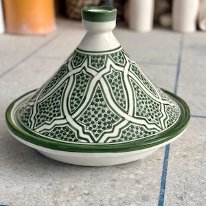 Peut inclure: Un tajine en céramique avec un couvercle conique présentant une base blanche et des motifs géométriques vert foncé complexes. Le couvercle possède un petit bouton au sommet, et l'ensemble est recouvert d'un vernis lisse et brillant.
