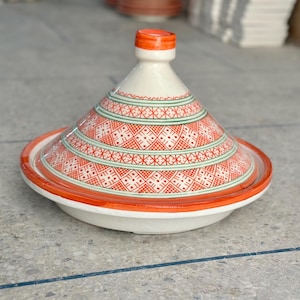Peut inclure: Un tajine en céramique avec un couvercle conique présentant une base blanche, un bord orange et des motifs géométriques rouges et verts complexes. Le couvercle possède un petit bouton au sommet et la base est un bol large et peu profond.