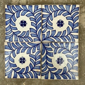 May include: Four square ceramic tiles with a repeating blue and white floral design. Each tile features a central white circle with a blue floral accent, surrounded by a blue leaf pattern. The tiles are arranged in a square.