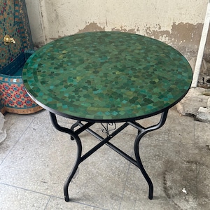Peut inclure: Une table ronde avec un plateau en mosaïque composé de petites tuiles vertes variées dans des tons émeraude, forêt et olive. La table est soutenue par un cadre en métal noir avec quatre pieds incurvés reliés par une structure en croix.