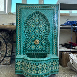 May include: A turquoise and gold mosaic tile fountain with an arched design. The fountain features a basin and a decorative spout. The intricate tilework includes geometric and floral patterns. The fountain is mounted on wheels.