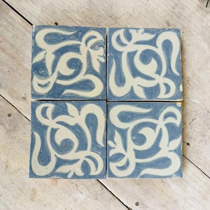 Könnte beinhalten: Vier quadratische Keramikfliesen mit einem blau-cremefarbenen Blumenmuster. Die Fliesen sind zusammen angeordnet und zeigen das sich wiederholende Muster. Die Fliesen liegen auf einer hellen Holzoberfläche.