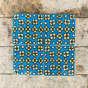 Puede incluir: Cuatro baldosas de cerámica cuadradas con un fondo azul vibrante y un patrón floral repetitivo. Las flores son amarillas con contornos negros y un centro verde. Puntos blancos están intercalados en el diseño.