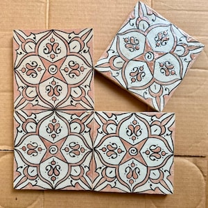 Peut inclure: Quatre carreaux de céramique carrés avec un motif floral rose, blanc et noir. Les carreaux sont disposés sur une surface en carton brun. Le motif floral présente une forme d'étoile centrale avec des détails ornés.