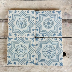 Può includere: Quattro piastrelle quadrate in ceramica con sfondo bianco e design a stella blu. Ogni piastrella presenta un motivo circolare centrale ed è decorata con piccoli accenti a forma di foglia. Le piastrelle sono disposte su una superficie di legno.