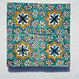Puede incluir: Cuatro baldosas de cerámica cuadradas con un patrón floral repetitivo. Cada baldosa presenta un diseño central de estrella de ocho puntas en amarillo, azul y blanco, rodeado de pequeñas flores blancas y azules sobre un fondo turquesa. Las baldosas están dispuestas en una cuadrícula de 2x2.
