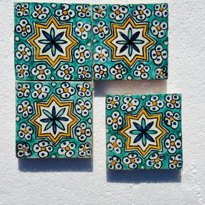 May include: Four square ceramic tiles with a turquoise background and a repeating floral pattern. Each tile features a central star design in yellow, white, and blue. The tiles are arranged on a white surface.