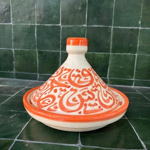 May include: A ceramic tagine pot with a cream-colored base and lid, accented with vibrant orange trim and calligraphy. The lid features intricate orange calligraphy. The pot is designed for cooking and serving Moroccan dishes.