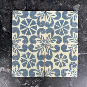Puede incluir: Cuatro baldosas de cerámica cuadradas con un diseño floral. Cada baldosa presenta un motivo floral central rodeado de hojas y espirales azules estilizadas sobre un fondo crema. Las baldosas están dispuestas juntas, mostrando el patrón repetitivo.