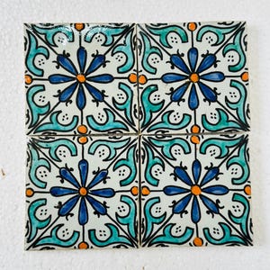 Puede incluir: Cuatro baldosas de cerámica cuadradas con un patrón floral repetido. Cada baldosa presenta una flor azul central con detalles en naranja, rodeada de elementos decorativos turquesa y negros. Las baldosas están dispuestas juntas, mostrando el diseño completo.