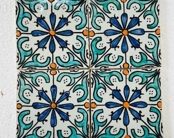 May include: Four square ceramic tiles with a repeating floral pattern. Each tile features a central blue flower with orange accents, surrounded by teal and black decorative elements. The tiles are arranged together, showcasing the complete design.