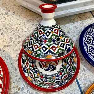 May include: A colorful ceramic tagine with a conical lid and matching base. The tagine features a vibrant geometric pattern in red, green, blue, yellow, and white. The base has a red rim and a detailed interior design.