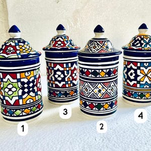 May include: Four ceramic jars with lids, showcasing detailed geometric patterns in red, blue, yellow, and green. The jars are cylindrical with a white base and a dark blue rim. Each jar has a unique design.