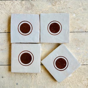 Peut inclure: Quatre carreaux de céramique carrés avec un fond gris clair et un motif de cercle marron foncé. Les carreaux sont disposés sur une surface en bois clair, présentant un motif géométrique simple. Le design est de style minimaliste et moderne.