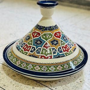 May include: A ceramic tagine pot with a conical lid and a round base. The pot features a white background with a colorful geometric pattern in red, blue, green, and yellow. The rim and lid are trimmed with a dark blue band.