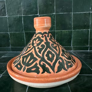 May include: A ceramic tagine pot, a traditional North African cooking vessel. It features a conical lid and a round base. The pot is orange with a black floral design on the lid. The base is white with an orange rim.
