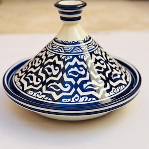 Peut inclure: Un tajine en céramique blanc et bleu, avec un couvercle conique et une base large et peu profonde. Le pot présente des motifs géométriques bleus et blancs complexes. Le couvercle a une petite poignée arrondie et est orné de rayures bleues.