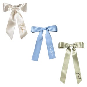 May include: Three satin bows in ivory, light blue, and champagne colors. Each bow has long, flowing tails and a name printed on one tail in a contrasting color. The ivory bow has "Nadine" printed in gold.