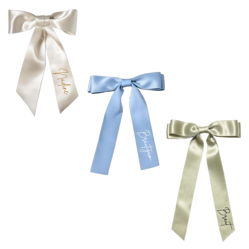 May include: Three satin bows in ivory, light blue, and champagne colors. Each bow has long, flowing tails and a name printed on one tail in a contrasting color. The ivory bow has "Nadine" printed in gold.