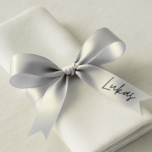 May include: A folded white linen napkin is adorned with a silver satin ribbon tied in a bow. The ribbon is printed with the name "Lukas" in a black script font, suitable for a wedding or special event.