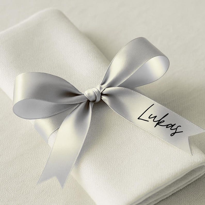 May include: A folded white linen napkin is adorned with a silver satin ribbon tied in a bow. The ribbon is printed with the name "Lukas" in a black script font, suitable for a wedding or special event.