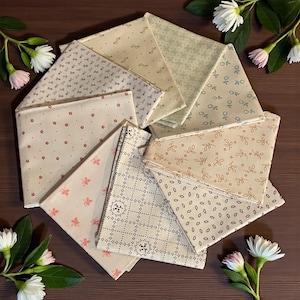 May include: A collection of folded fabric squares in various patterns and colors, including floral, dotted, and geometric designs. The fabrics are primarily cream, beige, and pastel green, with accents of red and pink. The arrangement is circular, with white flowers and greenery around the edges.
