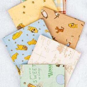 May include: A stack of colorful fabric squares featuring Winnie the Pooh and friends. Patterns include plaid, bees, and character illustrations in yellow, blue, and tan. The fabric is likely cotton and suitable for quilting or crafting.