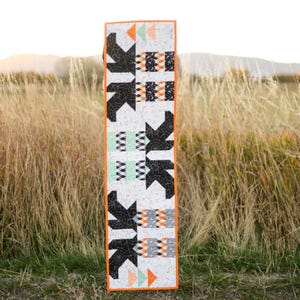 May include: A rectangular, vertically-oriented quilt with a white background and a repeating pattern of black arrow shapes, orange triangles, and geometric blocks. The quilt is bordered with orange fabric and is displayed outdoors in front of tall grass.
