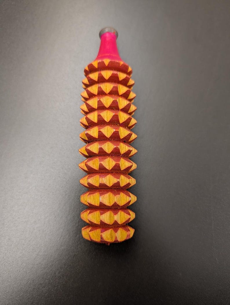 May include: A handcrafted wooden massage tool with a red and yellow geometric design. The tool has a cylindrical shape with a series of diamond-shaped protrusions. The top is painted red, and the base is a natural wood color.