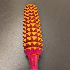 May include: A wooden massage tool with a textured, ridged head and a flared, pink handle. The head is composed of numerous yellow and red triangular shapes. The handle is pink with a green base.