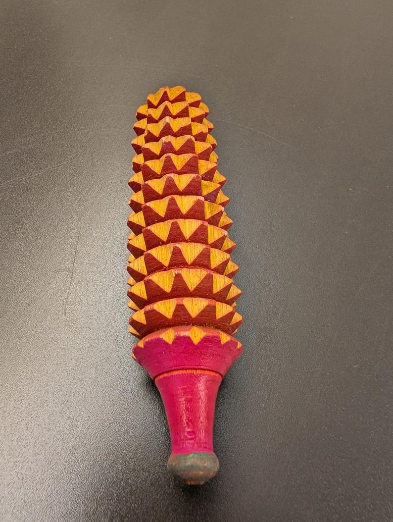 May include: A wooden massage tool with a textured, ridged head and a flared, pink handle. The head is composed of numerous yellow and red triangular shapes. The handle is pink with a green base.
